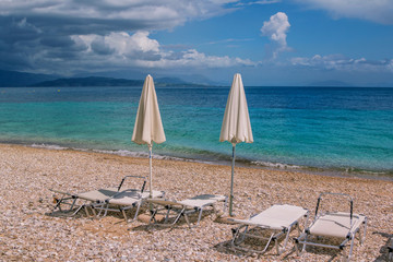 Fototapeta na wymiar View of empty beach – white deck chairs and umbrellas near sea water, pebbles and golden sand, mountains on the horizon and dark clouds on the sky. Landscape of abandoned summer resort. 