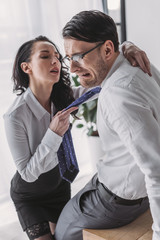 sexy secretary holding tie and hugging scared boyfriend while seducing him in office