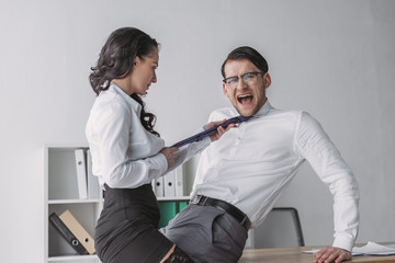 Obraz premium passionate businesswoman touching tie of shocked colleague while seductive him in office