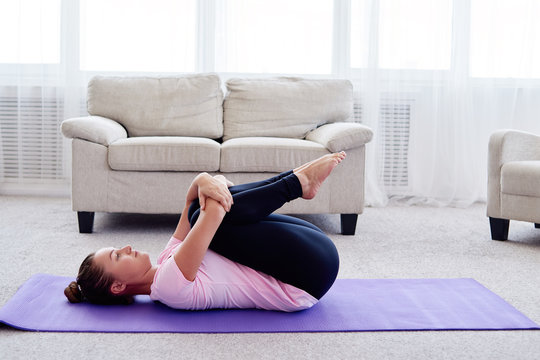 Portrait of young woman practicing apanasana pose on mat at home indoor, copy space. Apanasana. Knees to chest pose. Practicing yoga. Wellness and healthy lifestyle