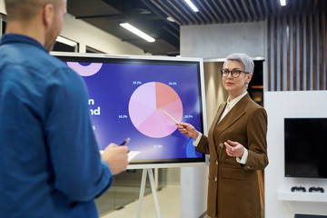 Portrait of modern mature businesswoman pointing at pie chart while giving presentation for employees or clients in office hall, copy space - Powered by Adobe