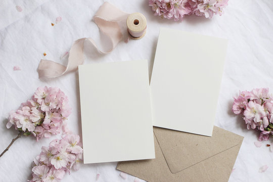 Wedding stationery mock-up scene. Blank greeting cards, envelope on linen tablecloth background with pink blossoming cherry tree branches and ribbon. Feminine still life composition. Flat lay,top view