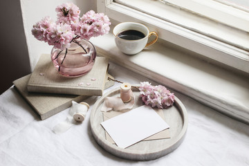 Spring still life scene. Greeting cards mockups, marble tray, cup of coffee, old books. Vintage feminine styled photo. Floral composition with pink sakura, cherry tree blossoms on table near window.