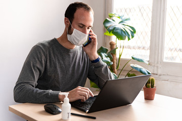 young man wearing medical face mask at work from home due to corona virus outbreak with laptop and...