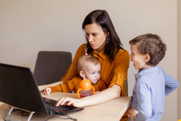 A young mother with two children works from home on a computer. Stress bad children behavior modern...