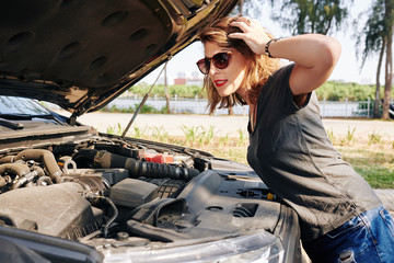 Stressed young woman opening car hood and looking at engine of broken car