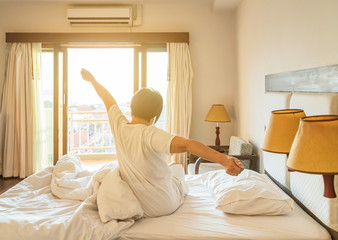 Back view woman stretching her arms in bed after wake up in the morning during sunshine shining through window with curtains in to the bed room, waking up concept
