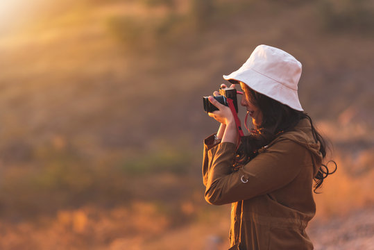 Happy smiling lifestyle portrait of pretty young woman traveler having fun while taking photos in evening on nature background. Beautiful girl photographer wear hat holding camera while travel at outd