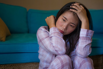 young beautiful sad and depressed Asian Chinese woman in pajamas at home sitting by living room couch feeling unhappy and confused suffering broken heart pain