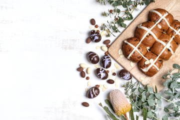 Chocolate Easter eggs and homemade hot cross buns on a wooden serve board surrounded by shaved almonds, hazelnuts plus Australian native Banksia and Eucalyptus leaves on a rustic white background.