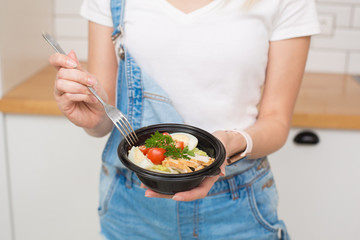 Happy girl in white shirt and blue jeans overalls, eats caesar salad at home, healthy food from delivery