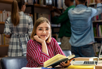 Young female student study in the library holding book in hand and looking at camera.