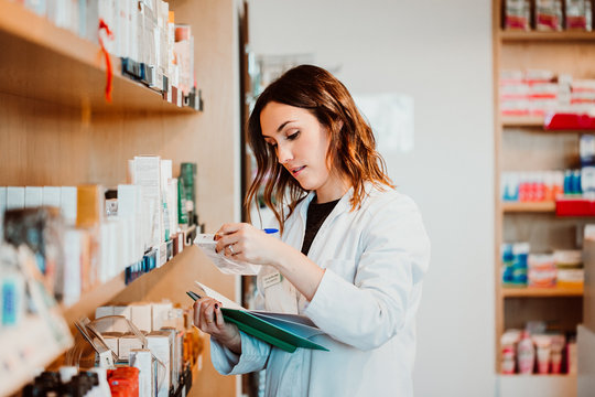 .Young female pharmacist working in her large pharmacy. Placing medications, taking inventory with her green notebook. Lifestyle