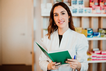 .Young female pharmacist working in her large pharmacy. Placing medications, taking inventory with her green notebook. Lifestyle