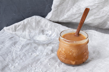 Glass jar with homemade salted caramel sauce on gray textile background.