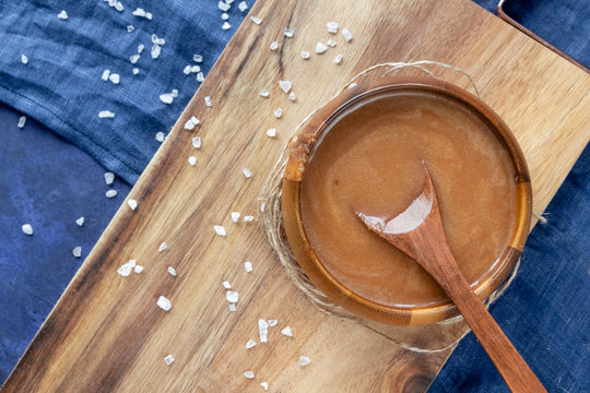  Homemade Salted Caramel Sauce In A Wooden Bowl On A Wooden Board With A Spoon, A Sea Salt And A Blue Textile Background. View From Above.