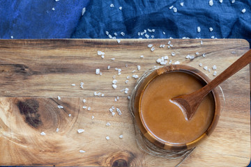  Homemade salted caramel sauce in a wooden bowl on a wooden board with a spoon, a milk, a sea salt and a blue textile background. View top.