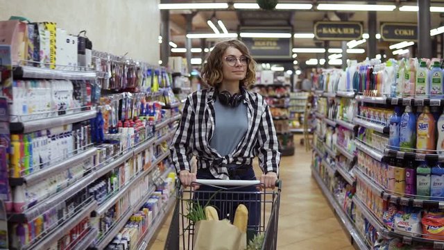 Modern young girl in glasses and headphones on neck walking with shopping cart in the supermarket and stearing around. Girl in plaid shirt and jeans. Slow motion