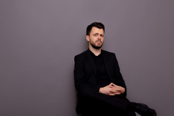 Obraz na płótnie Canvas Elegant man in a classic black suit sitting in studio against a grey background. Fashionably dressed serious businessman. Copy space.