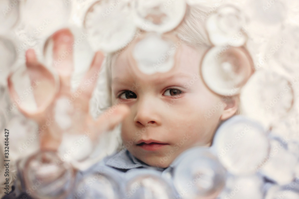 Sticker Portrait of blond toddler boy, lying under ice cubes, looking at camera