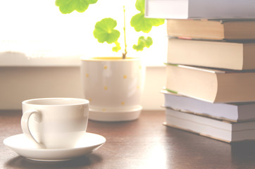 A white cup of coffee stands on a wooden table next to a stack of books and a pot against the background of the window