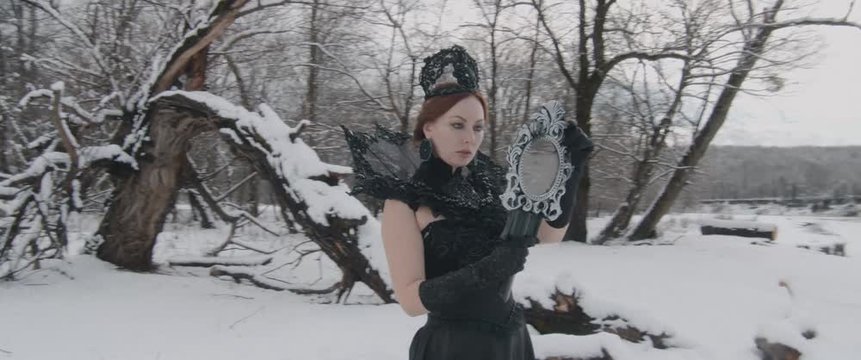 Charming model young woman in black long dress and crown on her head with mirror in fairy tale image stands in snow in winter fairy tale forest