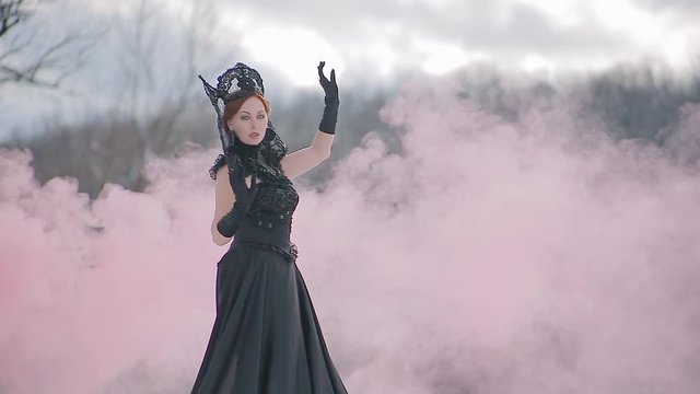 Charming model young woman in black long dress and crown on her head in fairy tale image stands in snow on background of winter forest and pink smoke