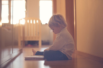 Beautiful toddler blond boy, lying on the floor at home in the hall, reading book