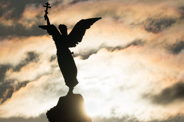Silhouette of an angel with a cross in his hand on a background of dramatic sky and sun. Fragment...