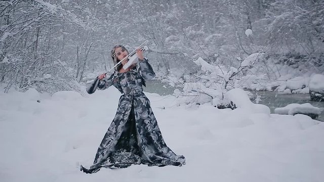 Amazing view charming cute slim young woman in fairy tale image in grey royal dress plays white violin on background of winter forest pictorial landscape with river with falling snow flakes