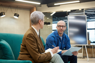 Obraz premium Portrait of bald mature man discussing work project with female manager while sitting on couch in modern office interior, copy space