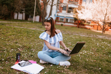  A young beautiful girl with a phone and a lap top is sitting on the grass in the park