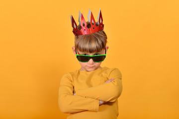 Glamorous and stylish boy model in sunglasses and with a crown on his head, in yellow clothes in the studio on a yellow background.