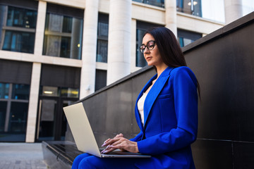 Serious woman executive director online banking via laptop computer while sitting outdoors near company building. Female government worker using notebook gadget for chat in internet