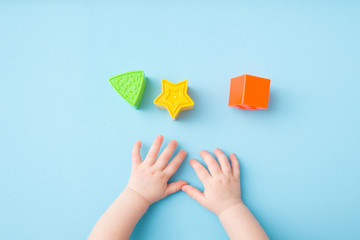 Baby hands playing with green triangle, yellow star and orange square shapes on light blue table background. Pastel color. Closeup. Toys of development for little kids. Top down view.
