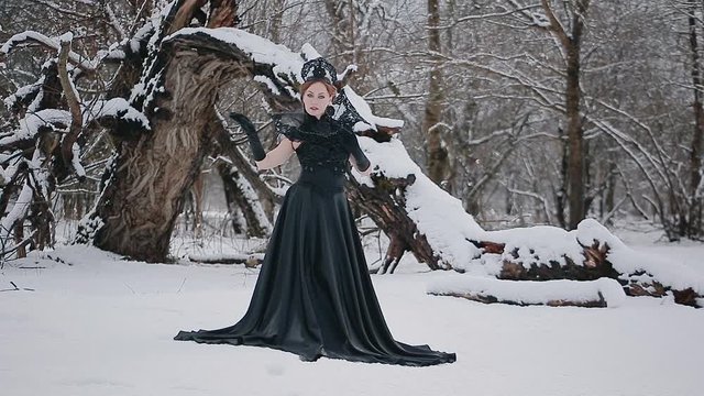 Charming model young woman in black long dress and crown on her head with mirror in fairy tale image stands in snow in winter fairy tale forest