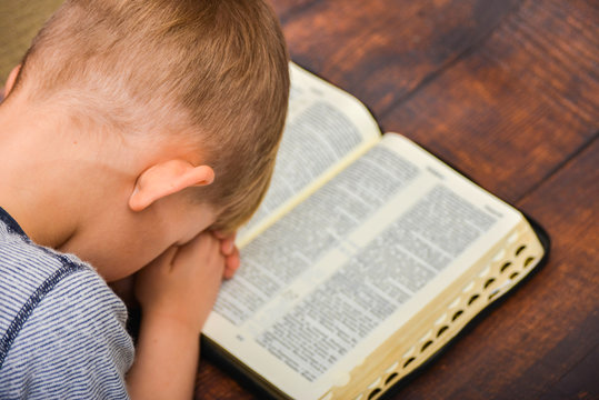 The Child Prays Over The Open Book Of The Bible, The Boy Folded His Arms And Bowed In Peace In Prayer To God.