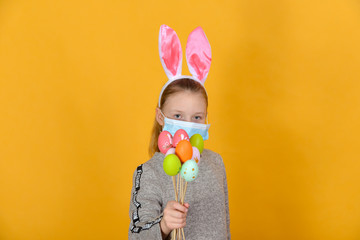 A girl with rabbit ears and a protective mask against coronavirus holds Easter eggs in her hand on a holiday.