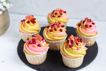 Traditional lemon muffins on a black tray and white background. With pink-yellow butter cream and pomegranate seeds.