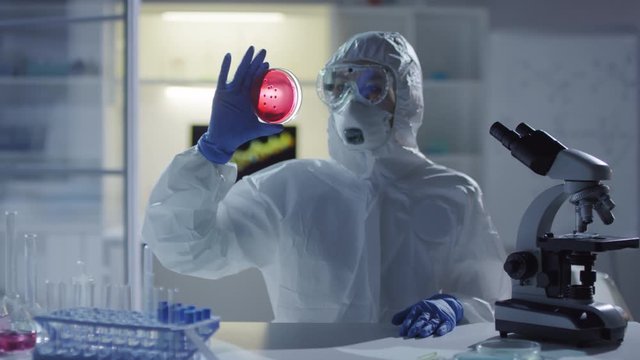 Dolly-in shot of unrecognizable medical scientist in disposable coveralls, goggles, respirator mask and gloves sitting at desk and looking at bacteria growing in petri dish