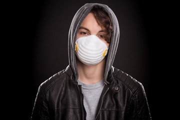 Concept of coronavirus quarantine. Teen boy wearing medical face mask to health protection from influenza virus, on black background. COVID-19 - home isolation. Emotional portrait of teenager in hood