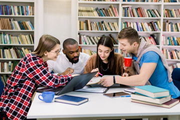 Young multiethnical college students at the library studying together, they are smiling, looking at book while finding new useful information for their exam or home task