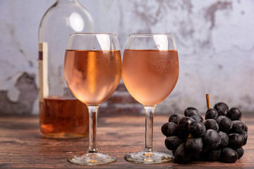 Two wine glasses full of pink/red wine with grapes laying next to it and a bottle of wine in the background on the wooden table with a broken concrete background