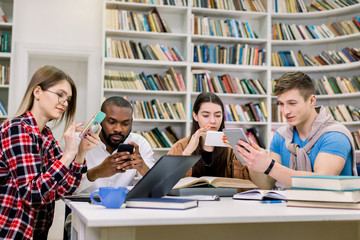 Four young friends students, boys and girls of mixed race, which are preparing for exams and searching needed information on their smartphones, sitting in the reading room in library