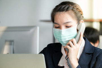 Women in the office wear mask to prevent corona virus. She pick up a phone and talk with customer and note into clipboard. Notebook and telephone on the desk. Protection virus and business concept.