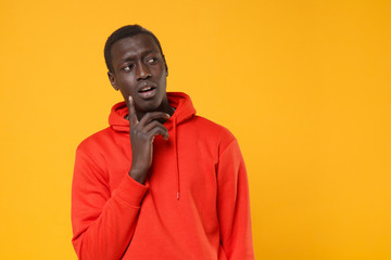 Pensive young african american man guy in red streetwear hoodie posing isolated on yellow background in studio. People lifestyle concept. Mock up copy space. Put hand prop up on chin, looking aside.