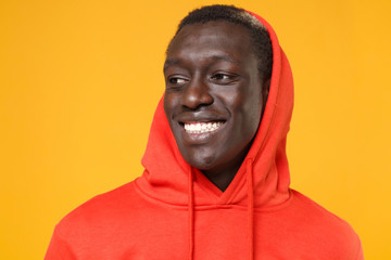 Close up of smiling young african american man guy in red streetwear hoodie with hood on head posing isolated on yellow wall background. People lifestyle concept. Mock up copy space. Looking aside.