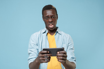 Irritated angry young african american man guy in casual shirt, yellow t-shirt posing isolated on blue background in studio. People lifestyle concept. Mock up copy space. Play game with mobile phone.
