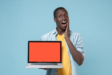 Excited young african american man guy in casual shirt yellow t-shirt posing isolated on blue background. People lifestyle concept. Hold laptop pc computer with blank empty screen put hand on cheek.