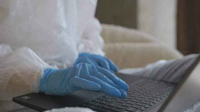 Woman working with laptop at home in a protective suit and gloves during the quarantine, close-up.